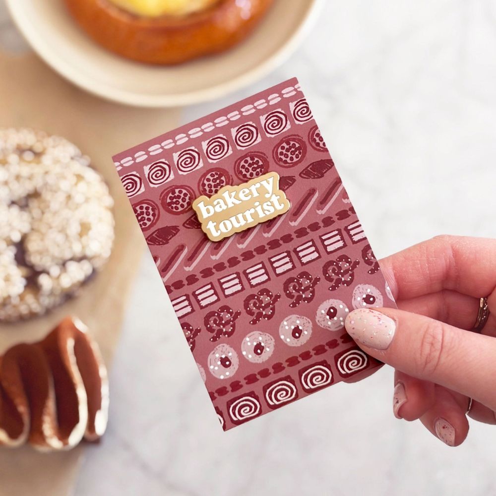 Hand holding a 'bakery tourist' pin badge mounted on a card with pastries in the background