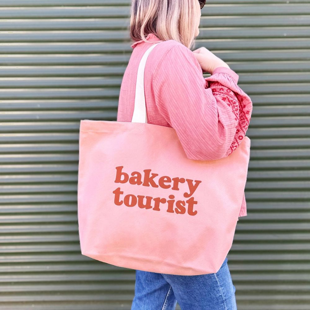 A person wearing a pink top and blue jeans carries a large light pink canvas tote bag with dark red text that reads 'bakery tourist' against a green corrugated metal background.