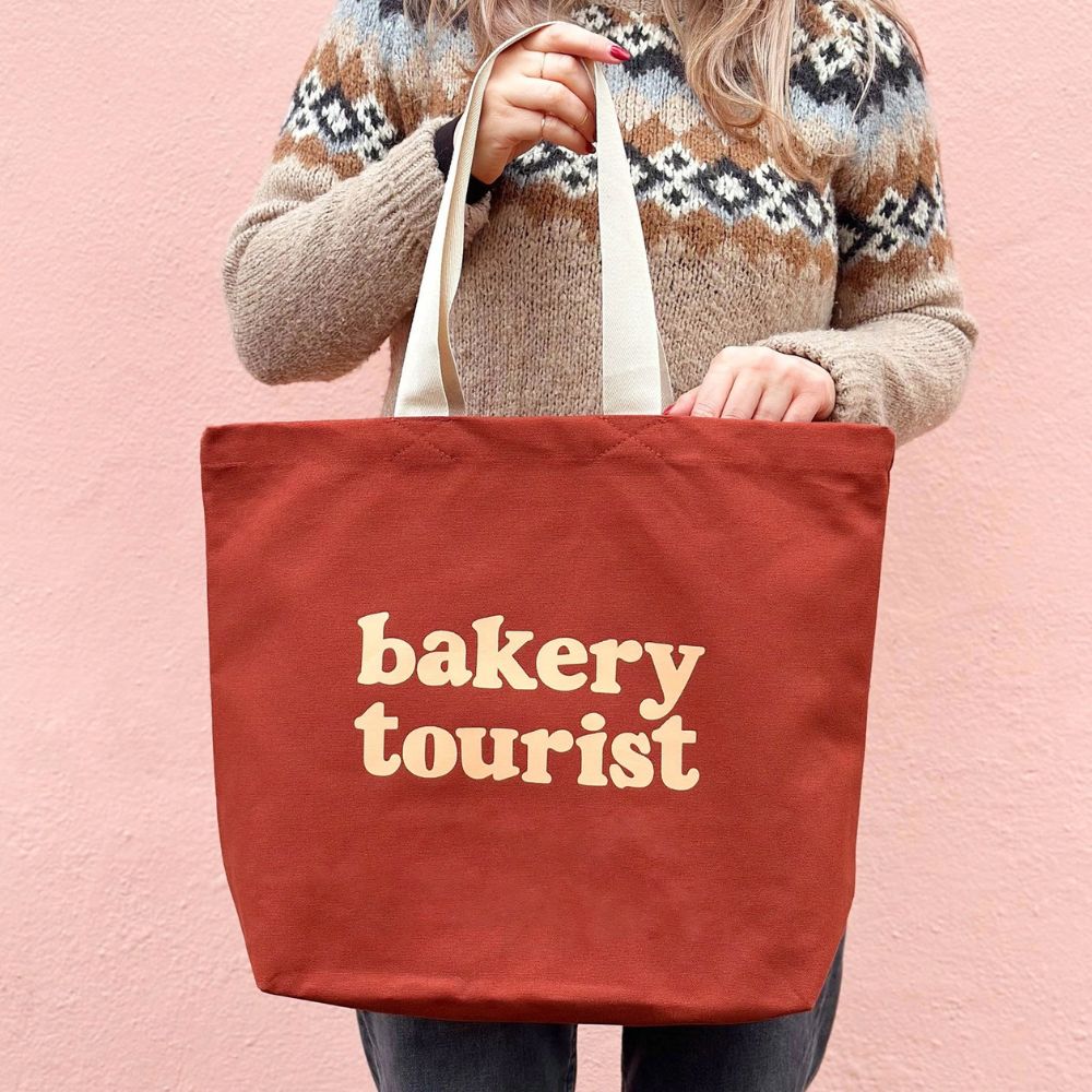 Woman holding a red-brown tote bag with the words 'bakery tourist' printed in large cream coloured letters on the front. The woman is wearing a nordic style knitted jumper and is stood against a pink wall