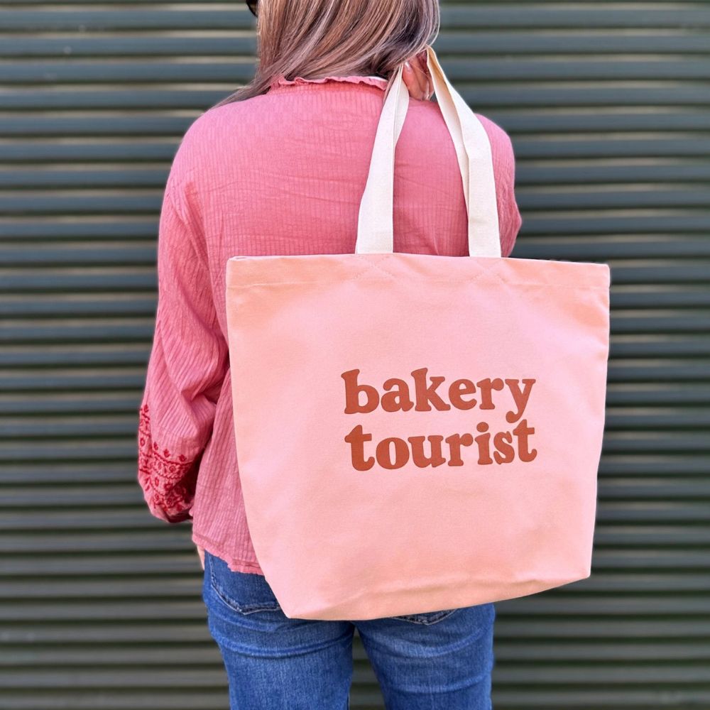 Person holding a tote bag with 'bakery tourist' text against a green metal shutter background