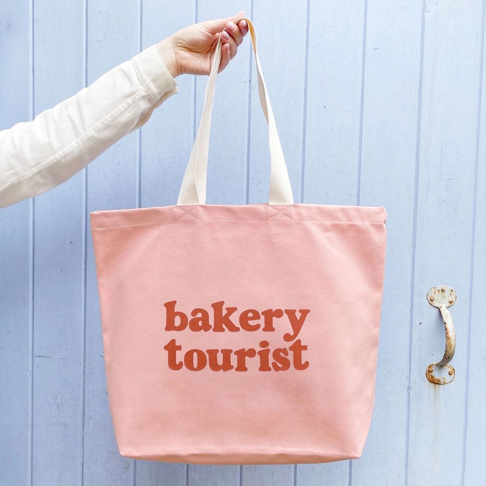 Pink tote bag with 'bakery tourist' text held by a person against a light blue wooden door.