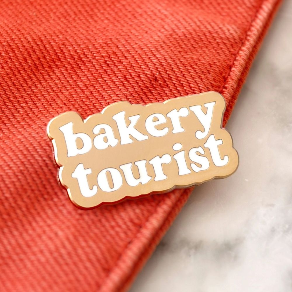A close-up of a scalloped enamel pin that reads 'bakery tourist' in white lettering on a gold coloured background, placed on a textured red-orange fabric surface with a light marble-like background visible at the corner.