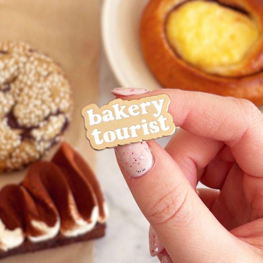 Hand holding a 'bakery tourist' badge with pastries in the background
