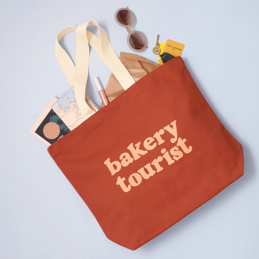 A terracotta orange canvas tote bag with cream straps and the words 'bakery tourist' printed in cream on the front, shown filled with sunglasses, a makeup compact, and other small items on a light blue background.