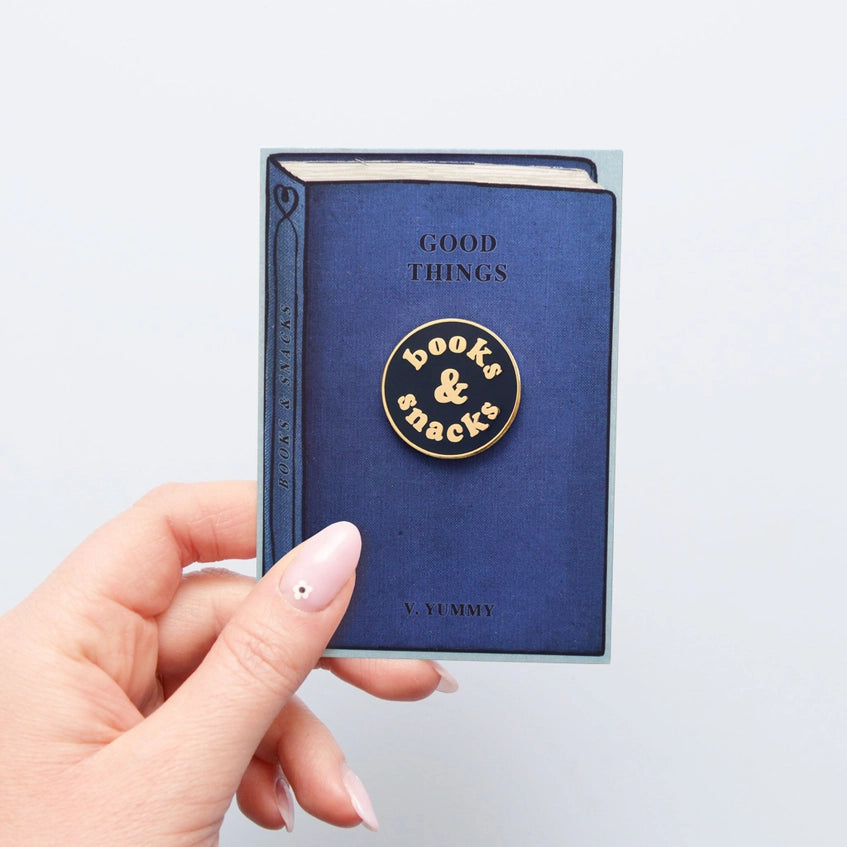 The pin badge being held on its backing card that's in the shape of a book.