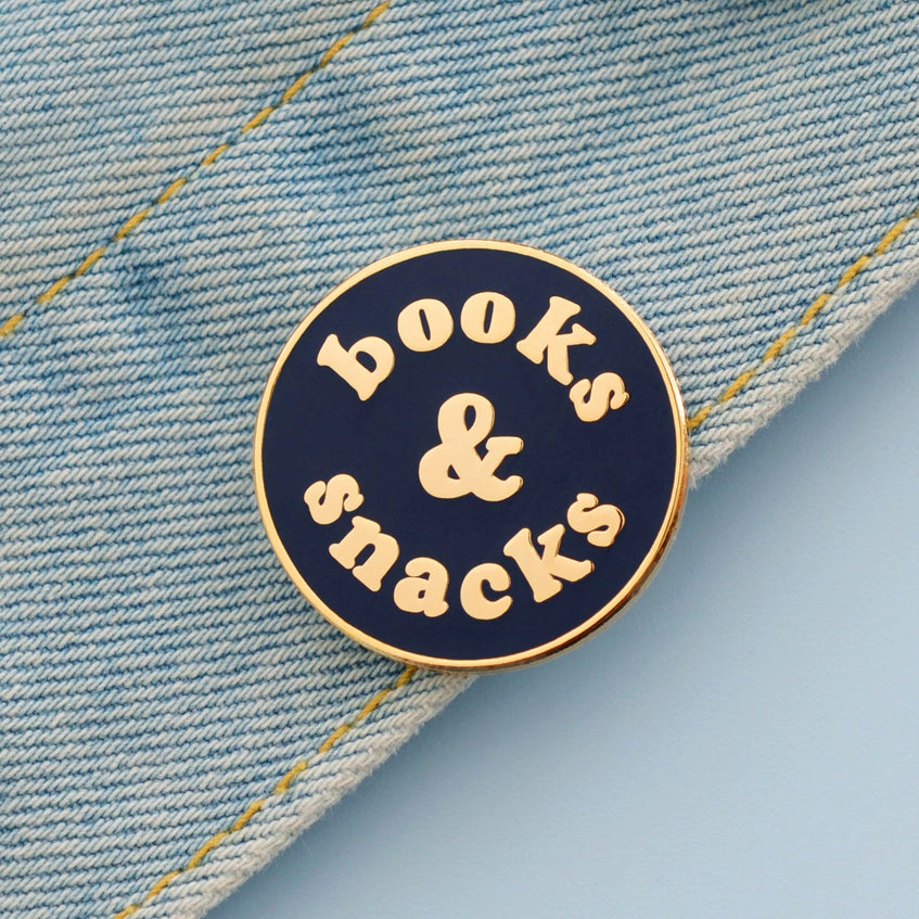 Books and snacks enamel pin badge shown on a light blue denim jacket, close up.