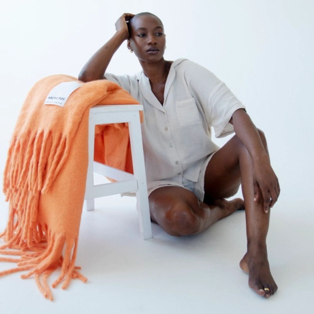 A woman leaning against a white wooden stool which the orange blanket is draped across